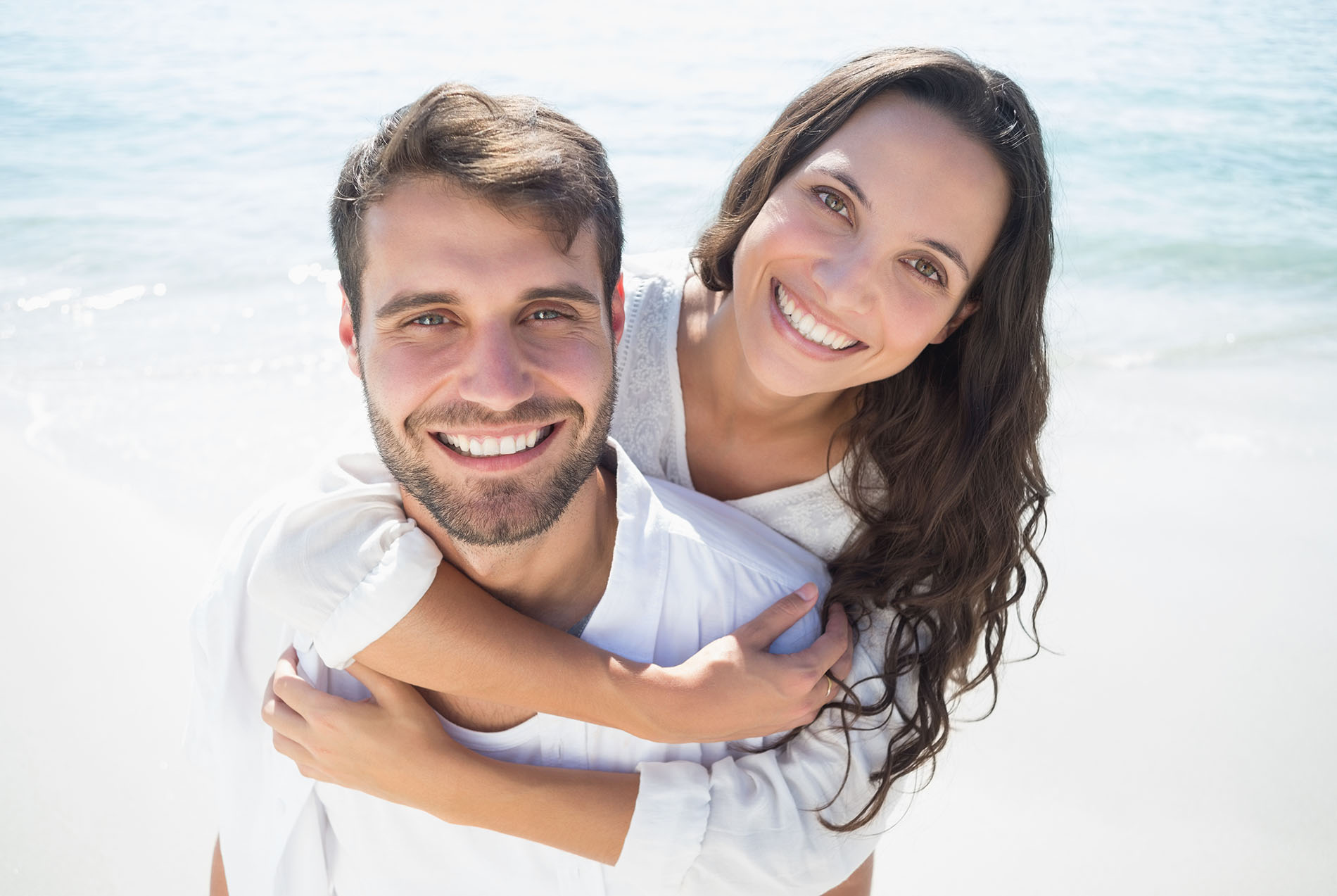 A man and woman are embracing each other on a beach with clear skies in the background.