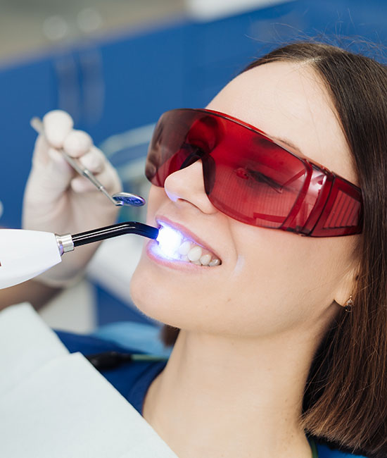 The image shows a person sitting in a dental chair with a dental professional performing a procedure, likely using a laser device, while wearing protective eyewear.